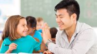 An elementary student and her teacher give each other a high-five at a table in a classroom.