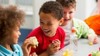 A closeup of two young boys and a young girl sitting at a cafeteria table, eating lunch, laughing and smiling together.