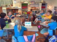 Group of eight elementary school children sitting in circle at desks, some with tablets