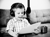 A black and white image of a young boy wearing headphones, reading from papers, speaking into a hanging microphone