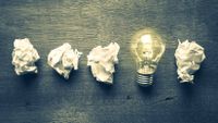 Four crumpled balls of paper and a lit light bulb illustrate the idea that success comes after failures.