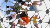 Eight young students are outside on top of a metal, geodesic dome at a playground, looking down. 