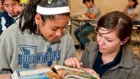 Girl sitting at her desk and an older girl kneeling to her left pointing to something in a book