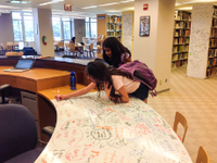 A photo of 2 female students writing a whiteboard table with colored markers.