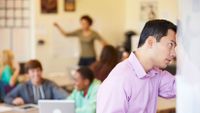 A male teacher is leaning his head against a whiteboard, looking up in distress. A classroom of blurred out students are behind him, smiling and talking.