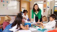 An elementary grade level teacher stands at a table giving instruction while young children are writing.