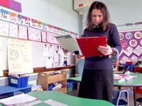 Teacher standing alone in classroom reading off a clipboard