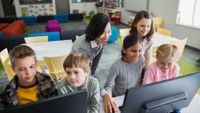 A teacher assists groups of students working at computers.
