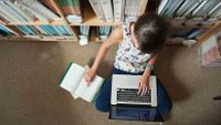 Photo shot from above of a young girl reading a book and typing on a laptop