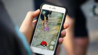 A person is holding a smartphone with Pokemon Go showing on their screen. They're taking a picture of someone bent over, holding a coffee cup, and touching a purple rat Pokemon.