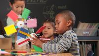 Three young, elementary-aged students are sitting at a classroom table building windmills out of cardboard and colorful popsicle sticks and styrofoam.