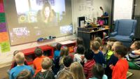 Children in a classroom talk to a writer who appears on a screen via video chat.