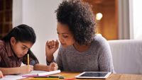 A woman is sitting at a wooden table with a young girl, helping her do her homework.