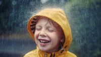 A young boy in a yellow rain coat is smiling with his eyes closed, and rain is pouring down on him.