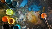 Paint brushes and jars of paint are on a black floor splattered with colorful paint.