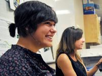 Two young women are sitting in a classroom, one smiling widely.