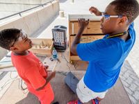 Two boys working with wood and circuits wearing goggles discussing a project