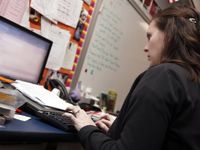 A side profile, closeup of a young, female teacher typing while looking at her laptop. She's sitting in her classroom toward the front of the room next to a whiteboard.