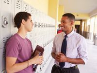 A male teacher and teenage boy are smiling, talking in a school hallway. The student is leaning against his locker, holding a folder and book.