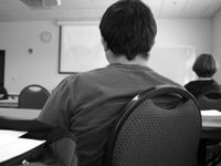 A black and white closeup of the back of a young man sitting at a long table towards the back of a classroom. He's looking toward the front of the classroom, and there's one other student sitting two tables ahead of him to the right.