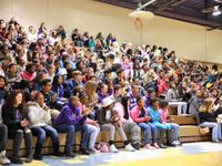 In the multipurpose room, a large group of middle school students fill one side of the bleachers, some clapping.
