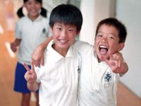 Two young, smiling boys standing in a school hallway wearing white polo shirts have an arm around each other's shoulder and are making the peace sign with their other hand.