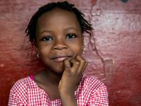 A closeup of a young girl, close-mouth smiling, with her chin resting on her right hand.