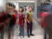 Two students are standing back to back in the middle of the school hallway. Other students are walking past them, blurred, as if to capture all the students walking through the hallway throughout the day.
