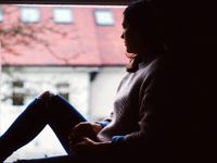 A young woman is sitting on a window sill in a dark room, looking outside the window.