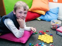 A young boy is lying on the floor, his elbows on a small, square pink pillow. He's looking at the camera with colorful shaped blocks in one hand, and more on the floor.
