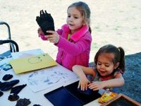 Two young girls are standing on grass next to a table with paper, stamp ink, crayons, and glove-like cloth that they're using as stamps. One girl is pushing her glove into the stamp ink, and the other girl is holding up her glove.