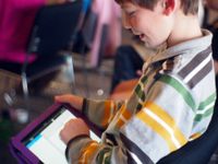 A closeup of the side of a young boy sitting in class, smiling, working on his tablet.