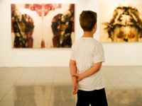 A young boy in a white t-shirt is standing in a museum, with his hands behind his back, looking at artwork.