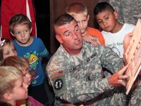 A U.S. army soldier in uniform is kneeling, reading to a group of young kids around him.