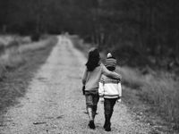 Two young kids are walking away from the camera along a gravel path with tall grass and trees on both sides. They're both wearing jeans and sweatshirts and one of them is wearing a beanie.