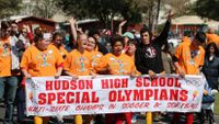 A large group of special needs students are marching in the street, proudly raising their arms, cheering, and holding a large sign that says, "Hudson High School Special Olympians -- Multi-State Champs in Soccer and Softball."