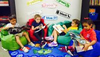 Three young girls and a boy are reading on bean bag chairs in class, each chair a different color: green, red, teal, and blue. Behind them are two bookshelves, and on the wall are mini posters with different colors written on them.