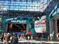 People are outside, walking towards a building that says "Come and Play," and "Welcome to Toy Fair NY 16."