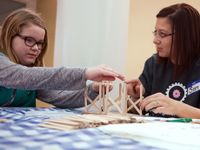 Two girls are sitting, building a house with popsicle sticks on a table with a white and blue checkered cover. 