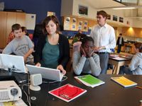 Three students are sitting at a long, black table in a large classroom. Two are on laptops, and one has her face in her hands with her elbows on the table. More students are sitting behind them at desks and a teacher is standing behind them.