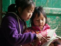 Two young girls, one in a purple sweatshirt and one in a pink sweatshirt, are sitting outside against a green, wooden door, reading together. 