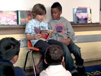 Two young students are sitting in chairs against the wall with a whiteboard behind them. They're reading a book to the rest of the class who are sitting on the carpeted floor in front of them.