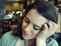 A closeup of a young women in a green shirt resting her head on her hand. The background, a coffee shop, is blurred.
