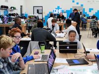 A classroom filled with high school students sitting at large rectangular tables, working together on laptops.