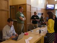 7 adults are surrounding two long tables pushed together. One is sitting, looking at his laptop. The rest are standing, some talking to each other, and some looking at their phones.