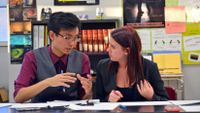 A teenage boy and girl dressed in business clothes are sitting at a classroom table -- covered with opened binders filled with paper -- talking to each other.