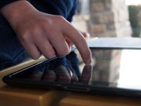 A closeup of a person's hand pressing their index finger against a tablet on a wooden table. In the upper right corner of the photo, there is a blurred corner of brick house and a tall green bush.