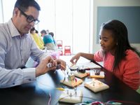 A female student and a male teacher are sitting at a raised table together putting wires together to turn on small light bulbs connected to square pieces of wood.