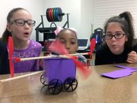 Three upper elementary girls are squatting next to a table, blowing on the wings of a toy car they've made, propelling it to move.