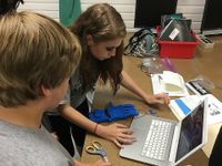 Three young teens are standing, hovering over a long, brown desk with folders, a red bin filled with notebooks, a pair of scissors, and opened folders with papers strewn across it. The girl is leaning towards a laptop, typing.